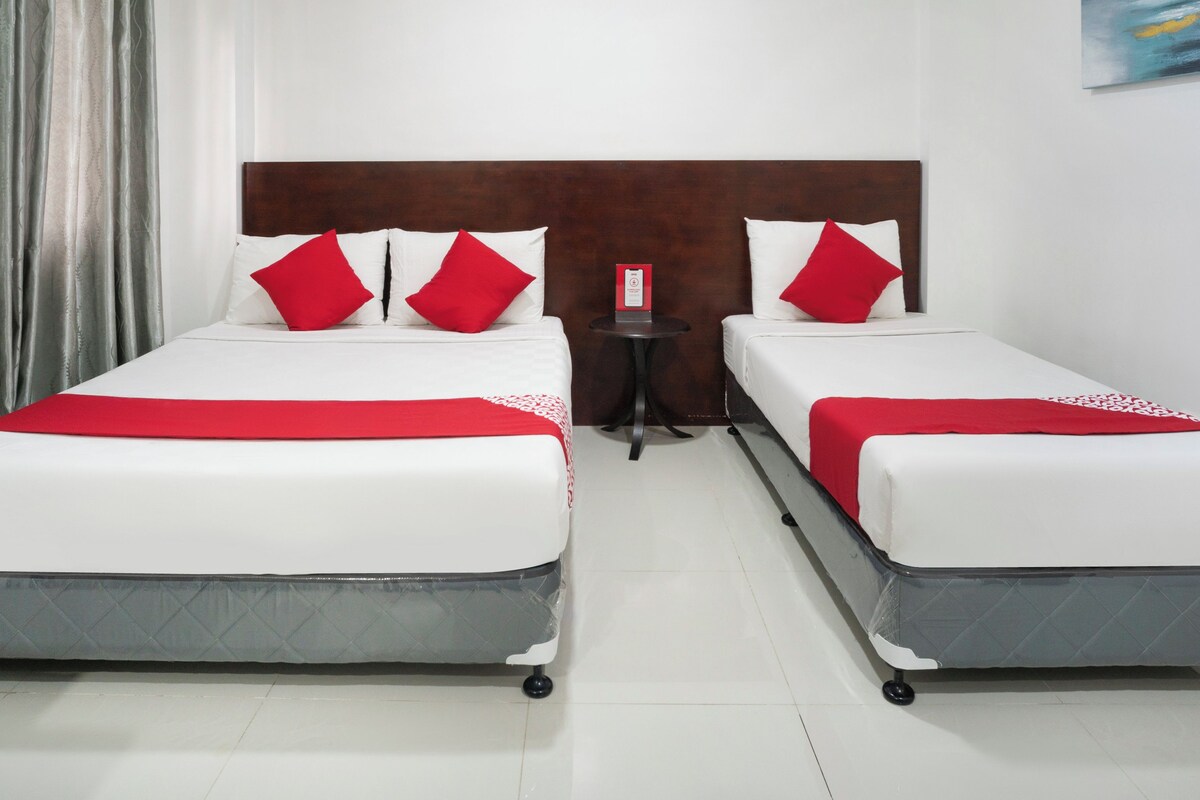 A hotel room with two beds, each with white bedding and red pillows. There is a small table with a red sign between the beds.