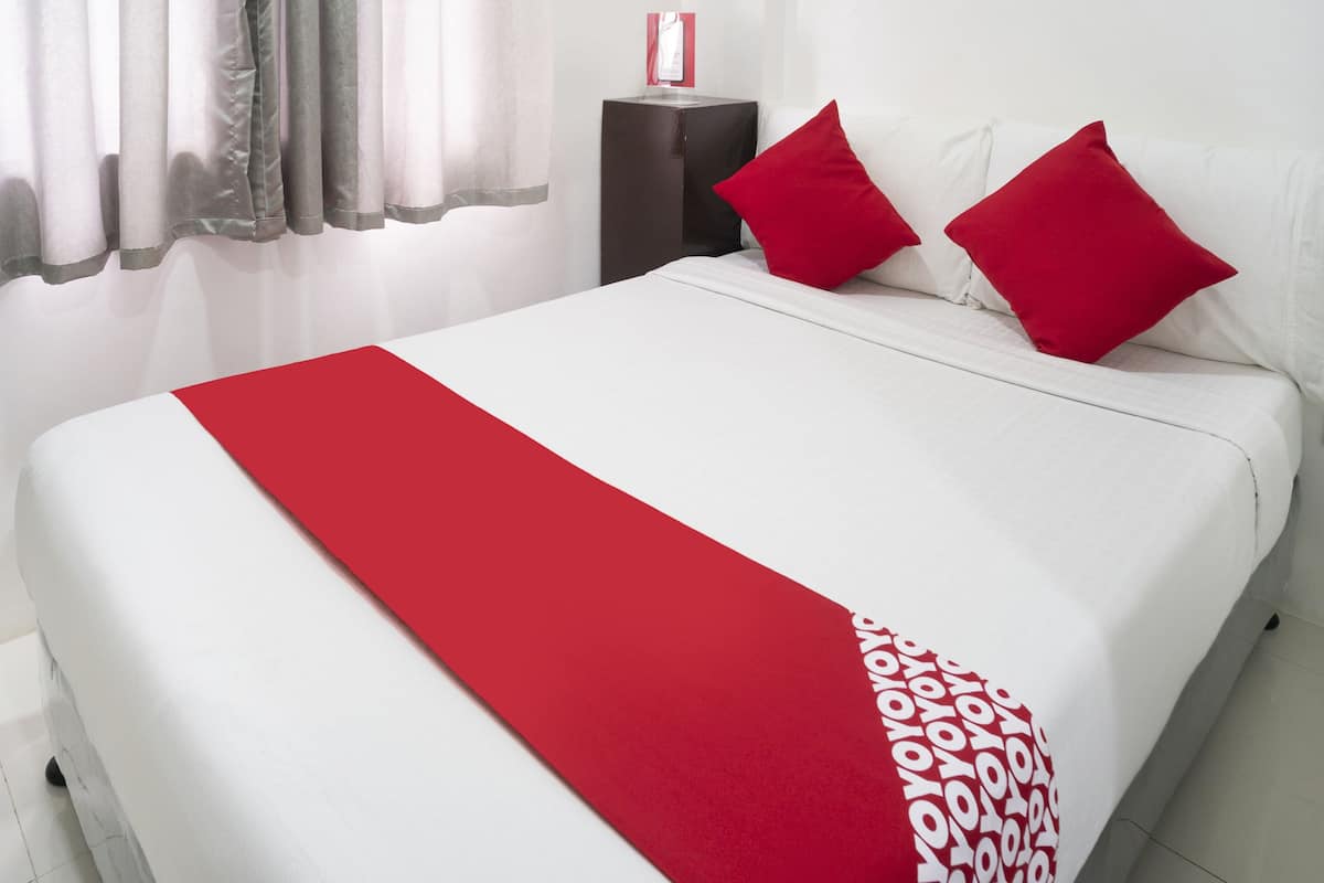 A neatly made bed with a red runner and two red pillows, a dark side table, and a white wall with a red object on it.