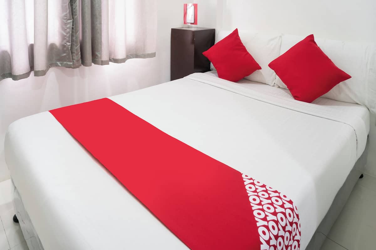 A neatly made bed with a red runner and two red pillows, a dark side table, and a white wall with a red object on it.