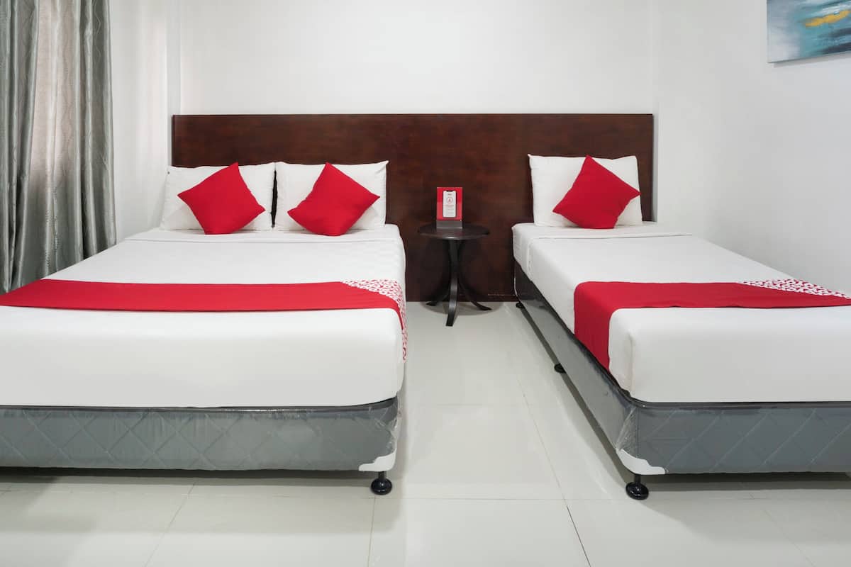 A hotel room with two beds, each with white bedding and red pillows. There is a small table with a red sign between the beds.
