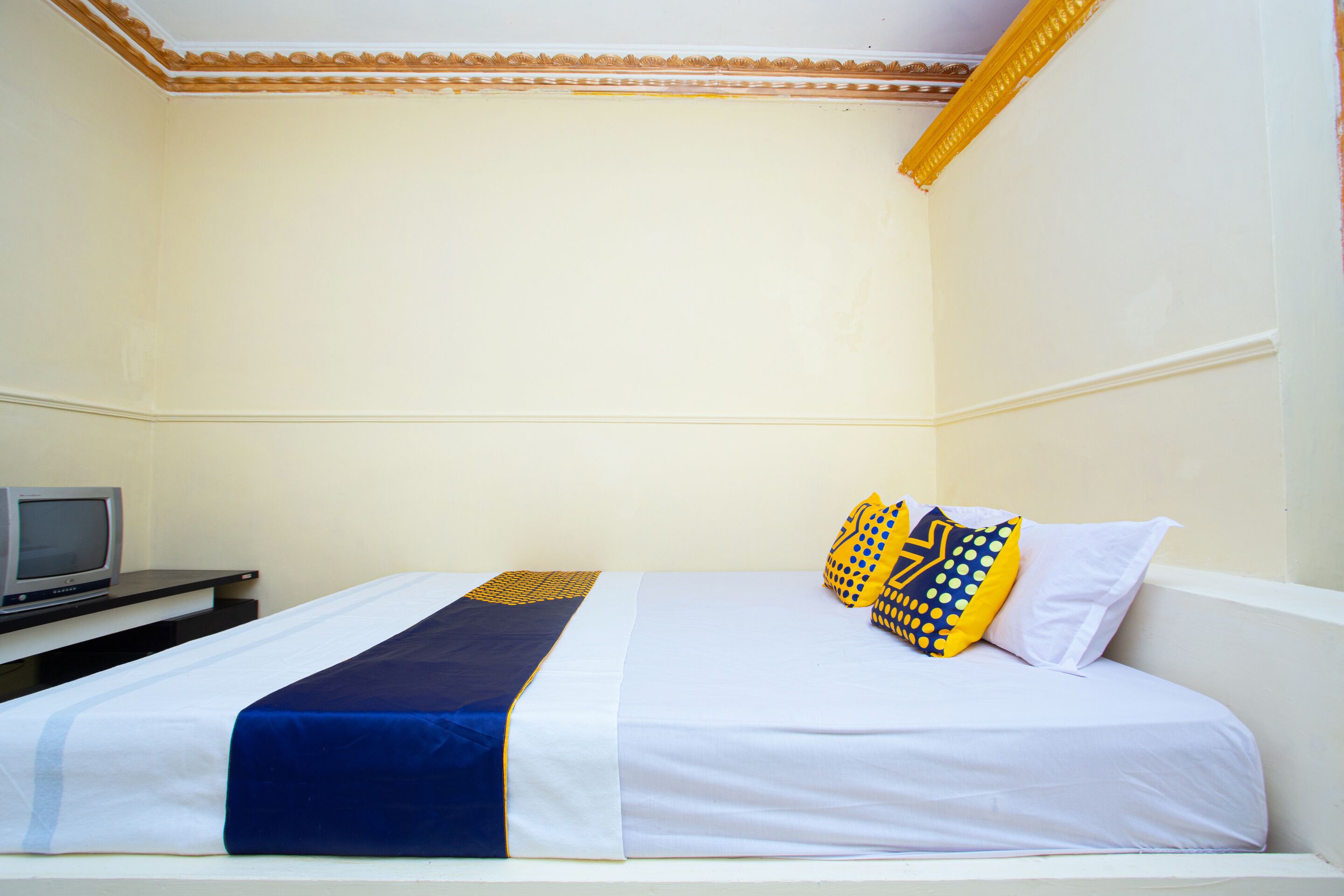 A neatly made bed with a blue and white bedspread, two pillows with yellow and blue patterns, and a television on a small table beside the bed.