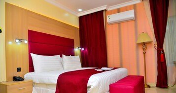 Habitación de hotel con una cama grande, cortinas rojas, aire acondicionado, una lámpara de noche y un teléfono junto a la cama.