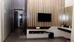 A modern bathroom with a large mirror, a flat-screen TV mounted on a wooden panel wall, and a built-in vanity with a sink and storage. - Hotel Central Park (Ambernath)