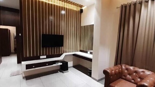 A modern living room with a flat-screen TV mounted on a wooden wall, a sofa, and a large window with curtains. - Hotel Central Park (Ambernath)