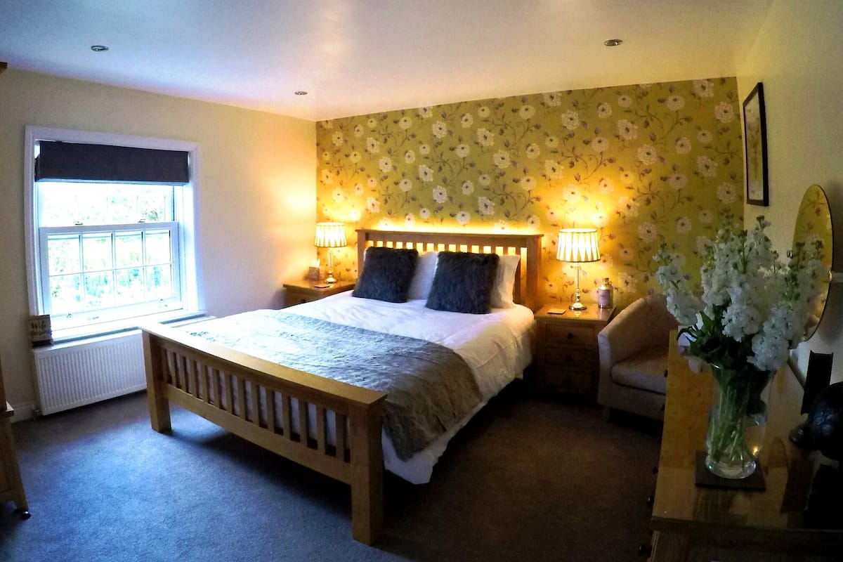 Quarto com cama de madeira, duas luminárias de cabeceira, mesa de cabeceira, uma cadeira, radiador, janela com persianas e papel de parede floral.