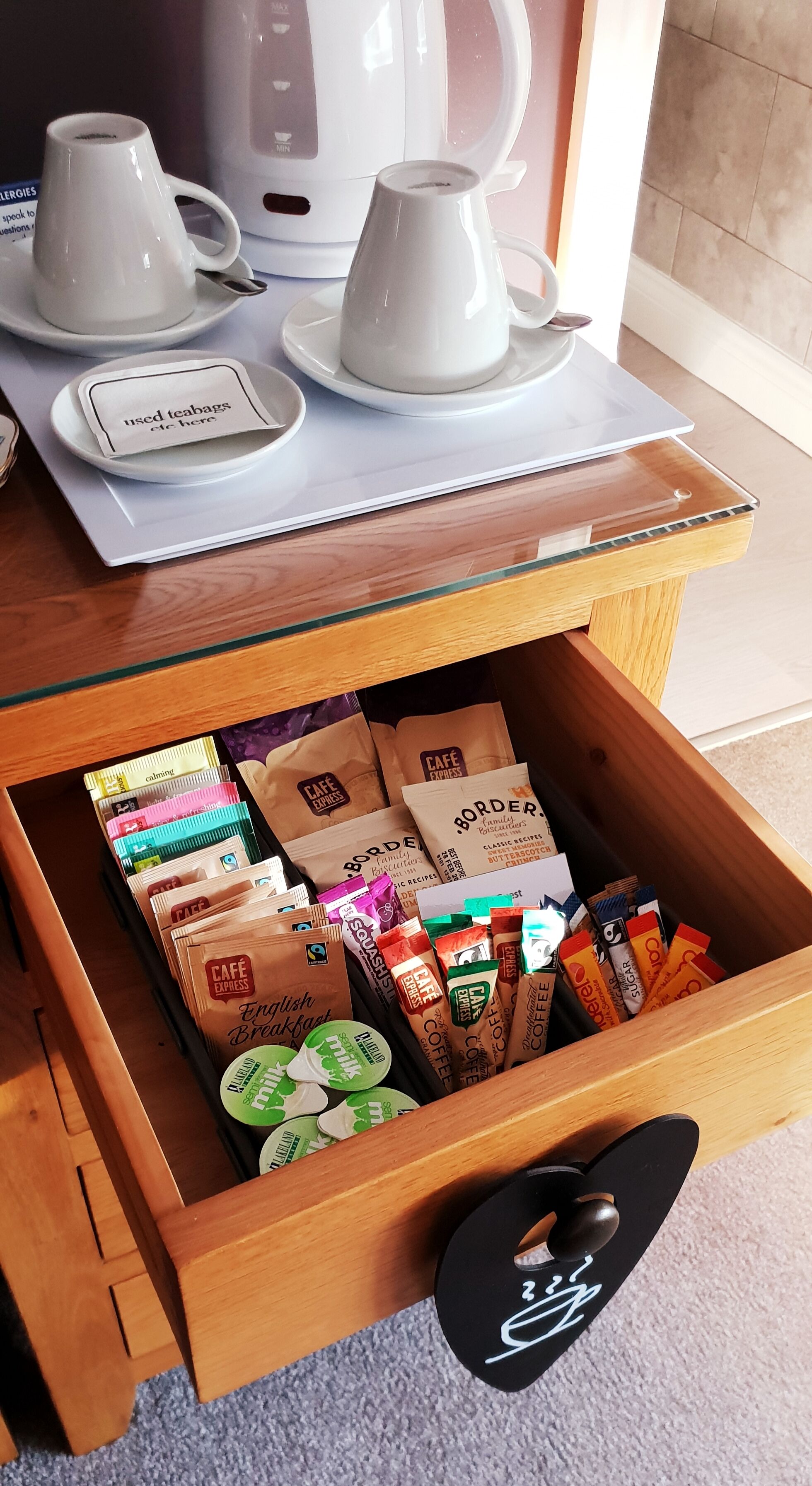 A coffee station with a coffee maker, two cups, and a tray with a teabag. Below, a drawer contains various coffee pods and a coffee bag.