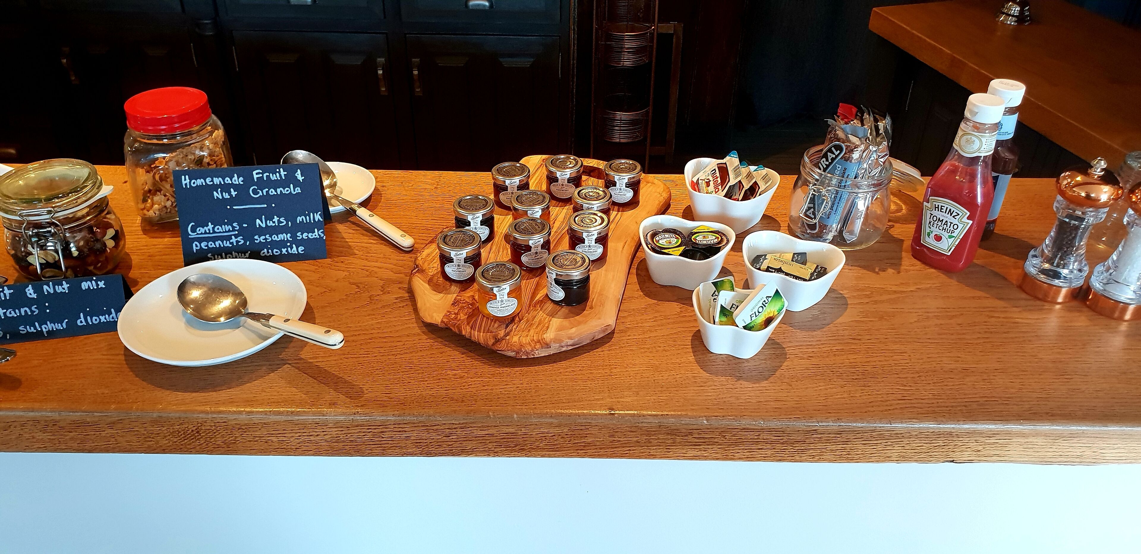 A display of homemade fruit & nut granola, condiments, and jars of preserves on a wooden counter.