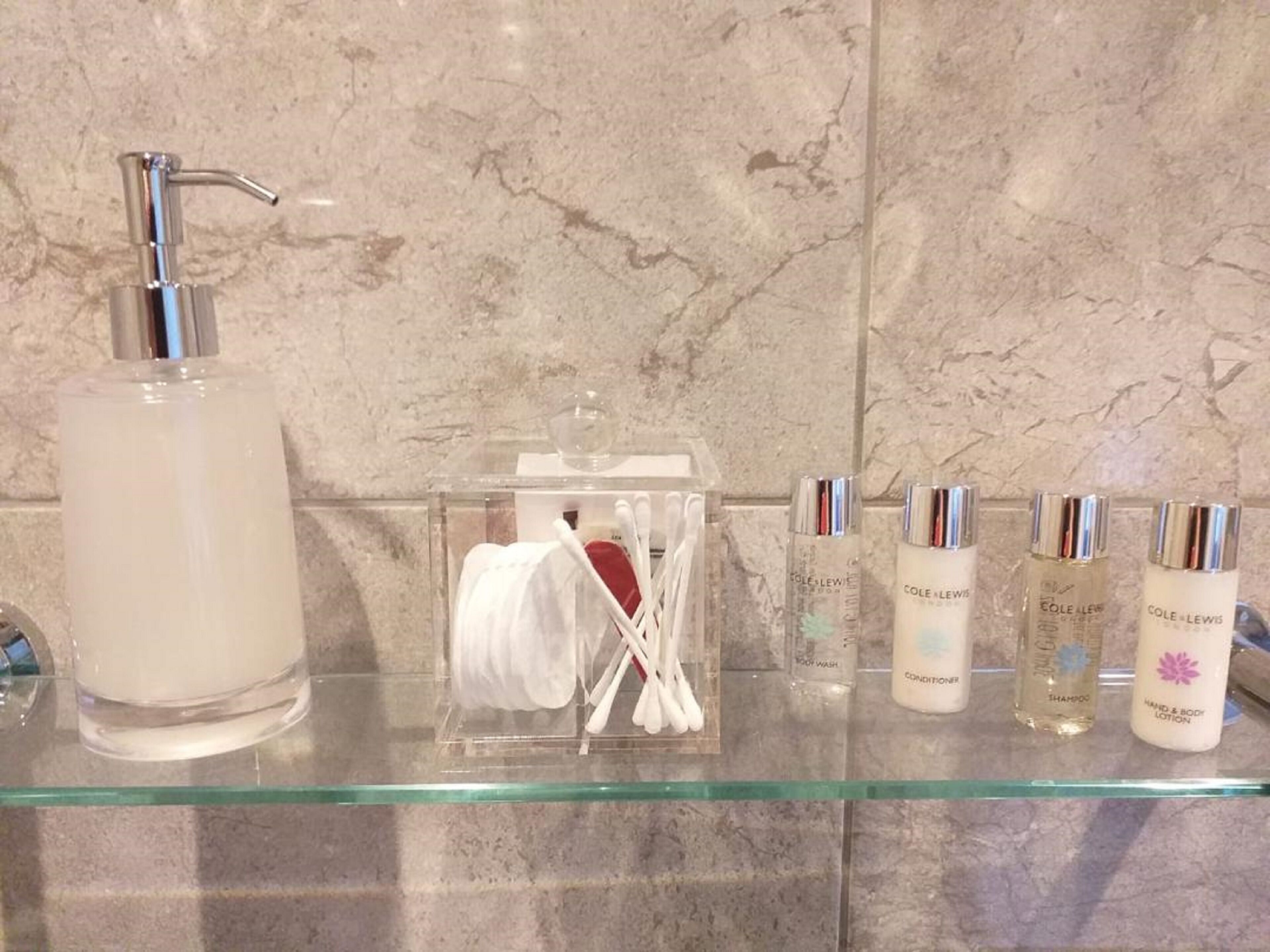 Un estante de baño con una botella de vidrio de jabón líquido, un dispensador de jabón transparente y varias botellas de productos de cuidado personal, incluyendo champú, acondicionador y loción.