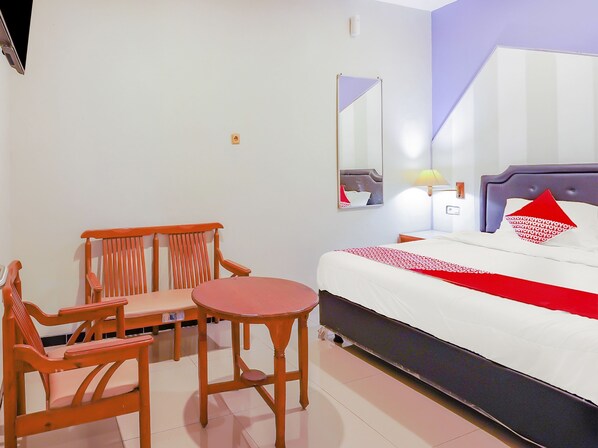 Habitación de hotel con cama, una silla, una mesita, un espejo y una mesita de noche con lámpara.