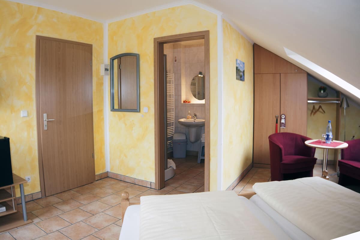 A hotel room with a bed, a desk, a chair, a small table with a bottle, and a mirror on the wall.