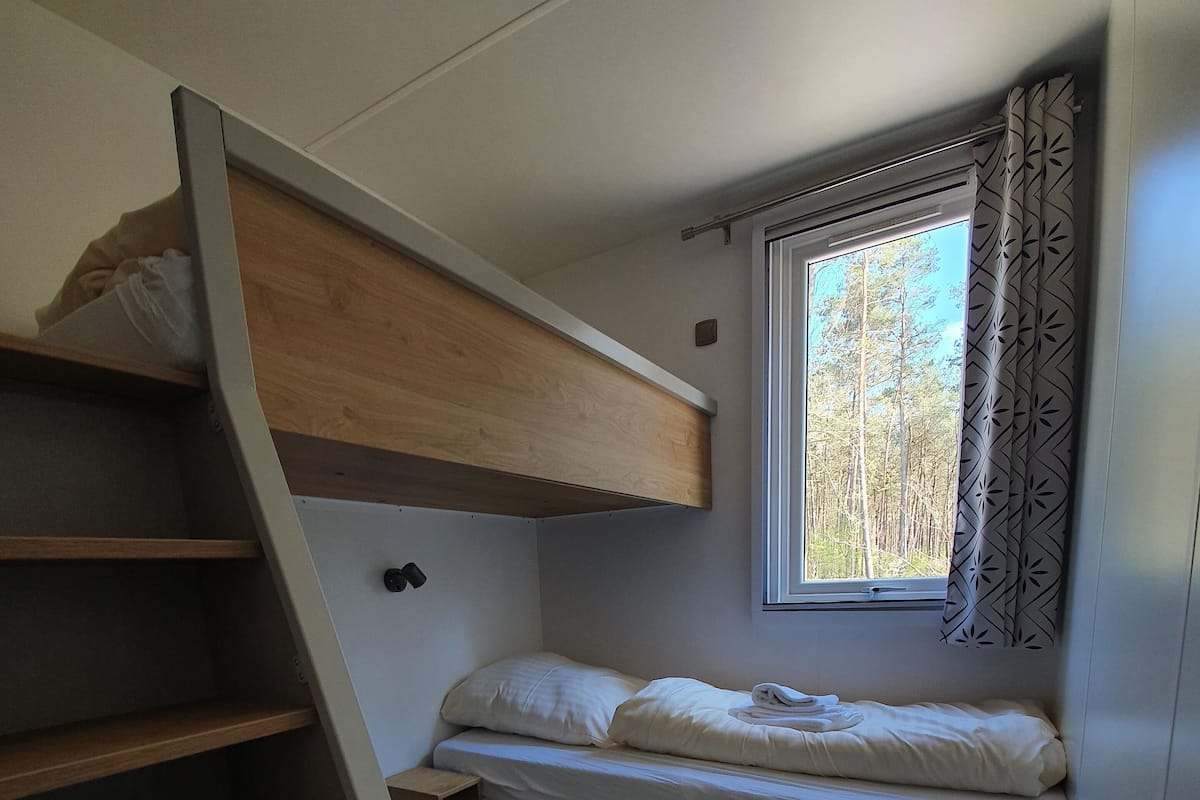 A compact room with a bunk bed, a small bedside table, and a window with curtains.