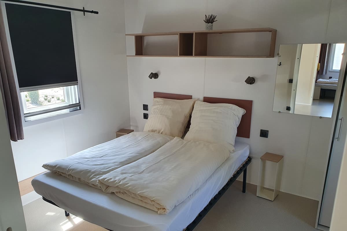 A neatly made bed with a headboard, a bedside table, a small shelf, and a window with curtains.