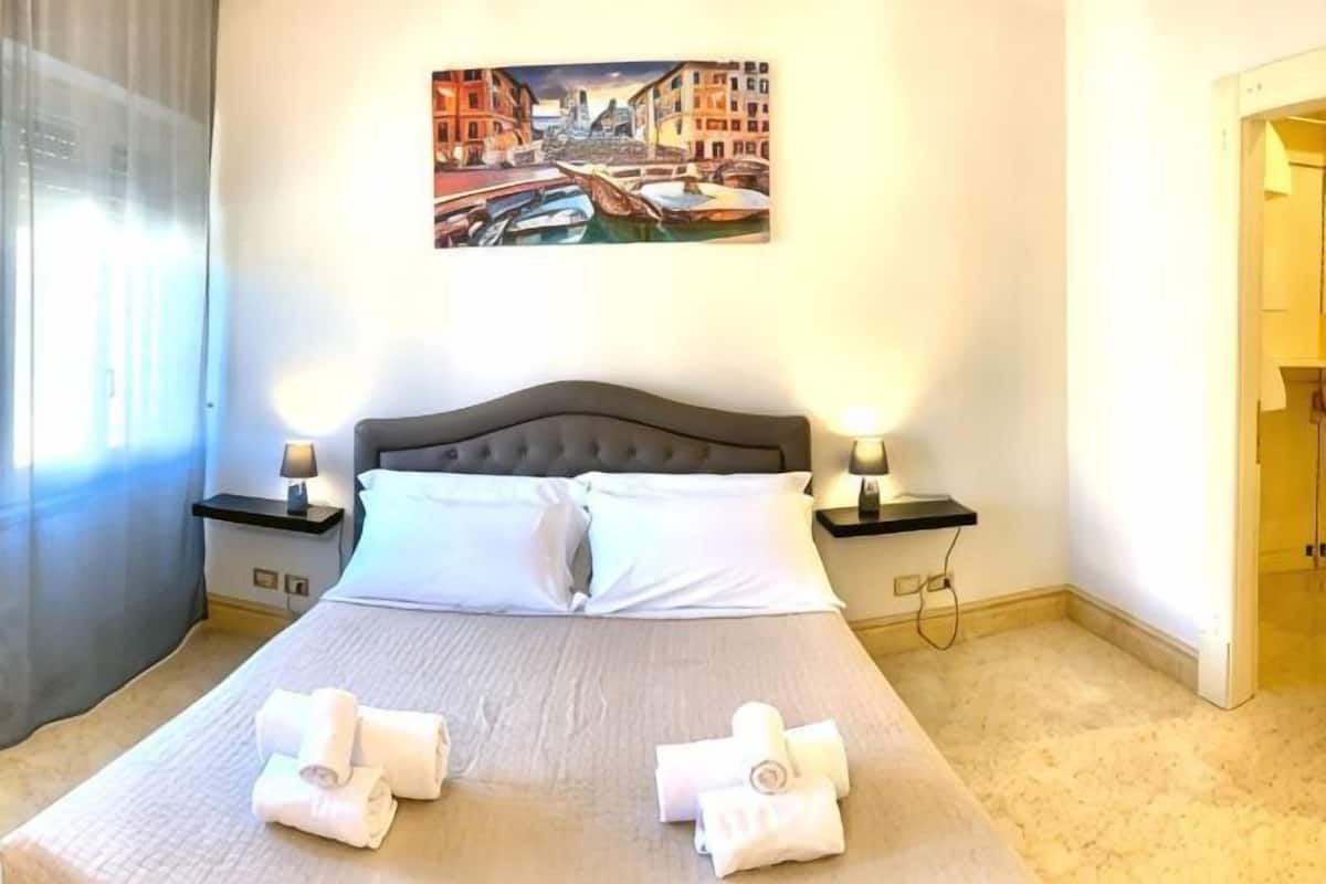 A neatly made bed with two pillows, a painting of a cityscape on the wall, and two bedside lamps.