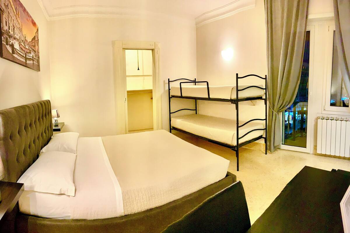 A hotel room with a large bed, a nightstand, a lamp, a bunk bed, a window with curtains, and a radiator.
