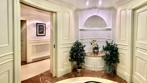 A hallway with cream-colored walls, a marble floor, and a framed picture on the wall. There are two potted plants on either side of the hallway. - Simon's Guest House Rome (Rome)
