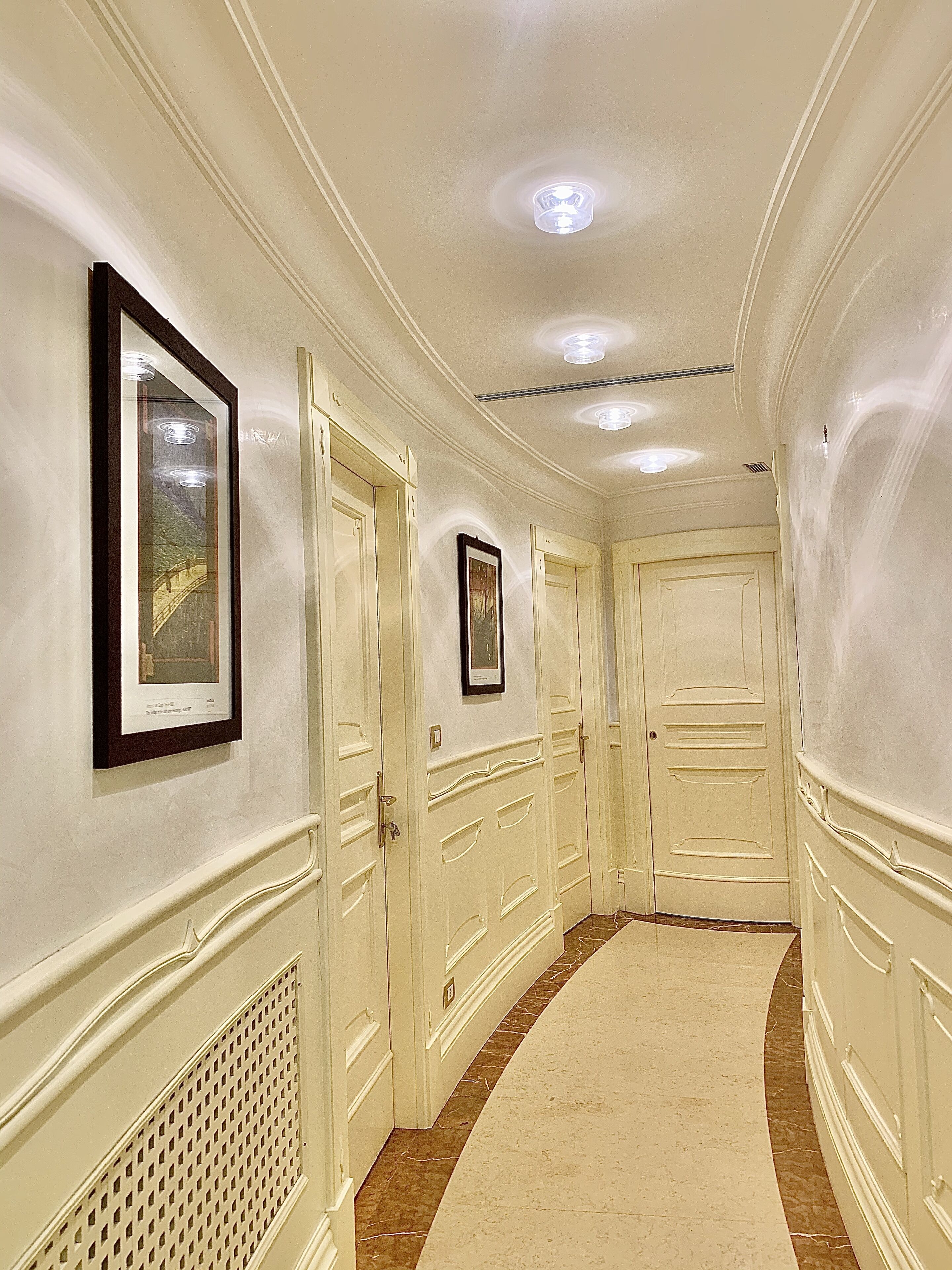 A hallway with cream-colored walls, white doors, and a marble floor. There are framed pictures on the walls and a ceiling light fixture.