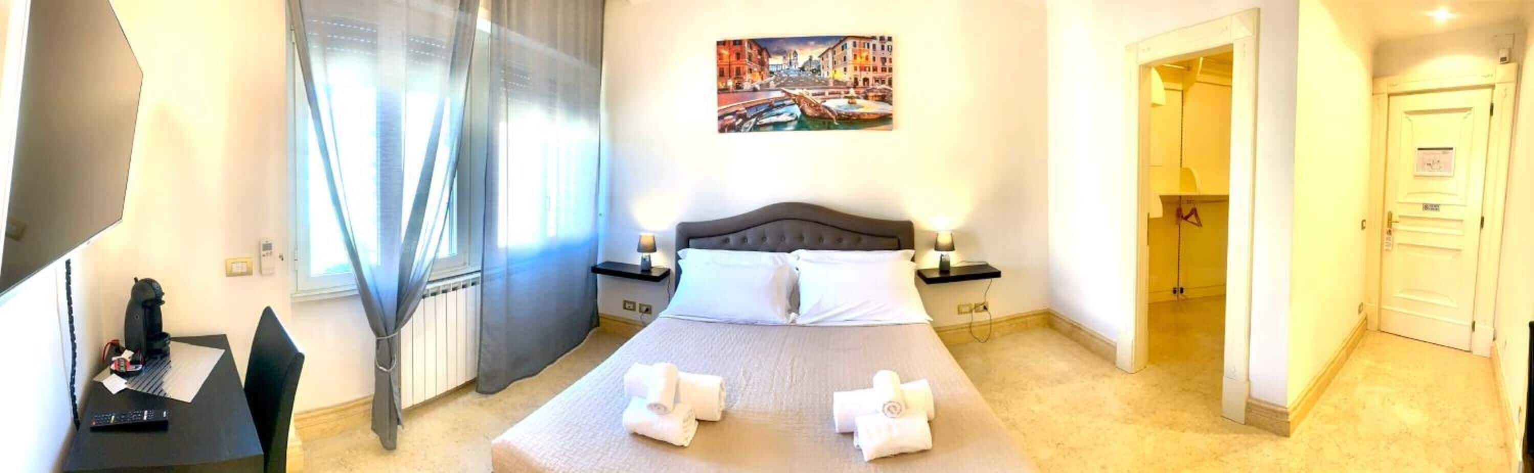 Une chambre d’hôtel avec un lit, un bureau et une chaise, une fenêtre avec des rideaux, une table de chevet avec une lampe et un tableau au mur.