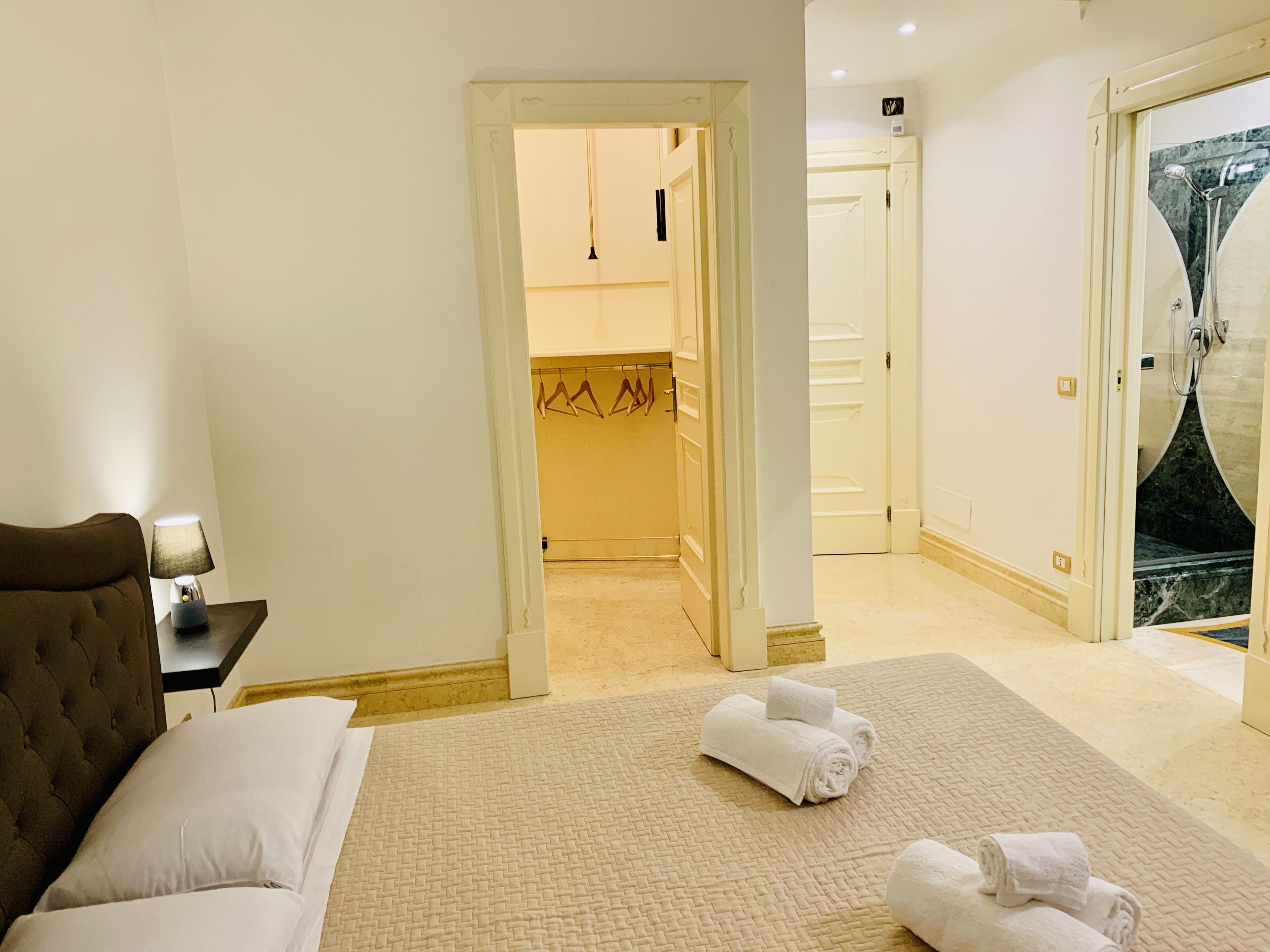 A hotel room with a bed, a nightstand, a lamp, a bathroom with a shower, and a door leading to a hallway.