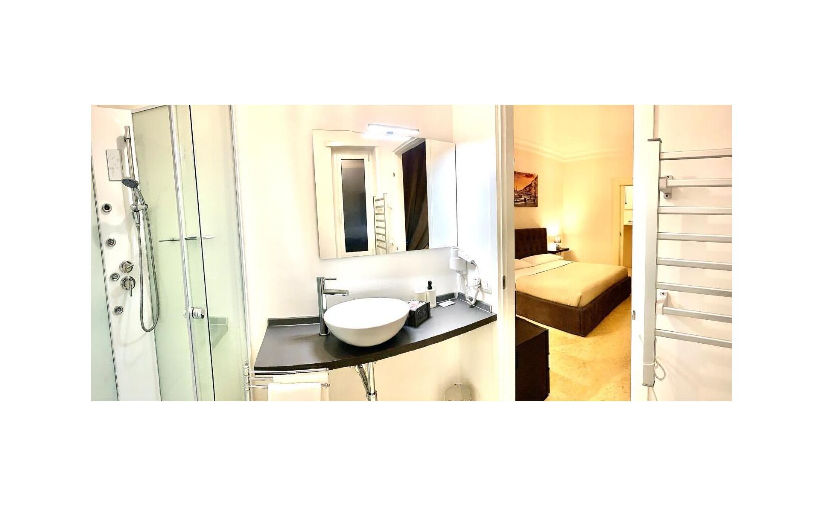 A bathroom with a glass shower enclosure, a white sink with a mirror above, and a bed with a headboard and bedside table.