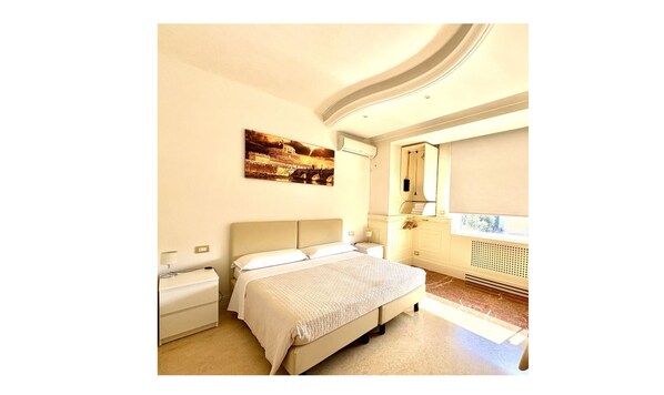 Un dormitorio con cama, mesitas de noche, aire acondicionado instalado en la pared, una ventana con persiana y un cuadro de un puente.