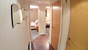 Un couloir au plancher de bois menant à une chambre à coucher équipée d’un lit et d’une table de chevet.