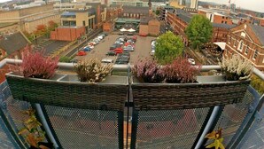 Une vue depuis un balcon donnant sur un parking où l’on aperçoit différentes voitures, des bâtiments et, au loin, des grues de construction.