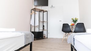 Habitación de hotel con dos camas, un televisor montado en la pared, un escritorio con dos sillas y un armario con ropa colgada.