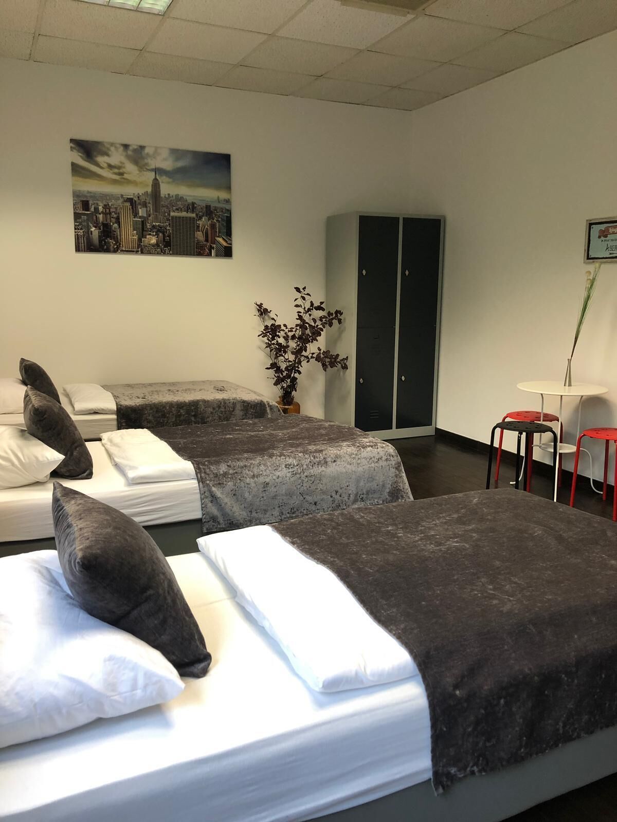 Ein Hotelzimmer mit zwei Betten, einem Stadtbildgemälde, einer Topfpflanze, einem Kleiderschrank und einem kleinen Tisch mit Stühlen.