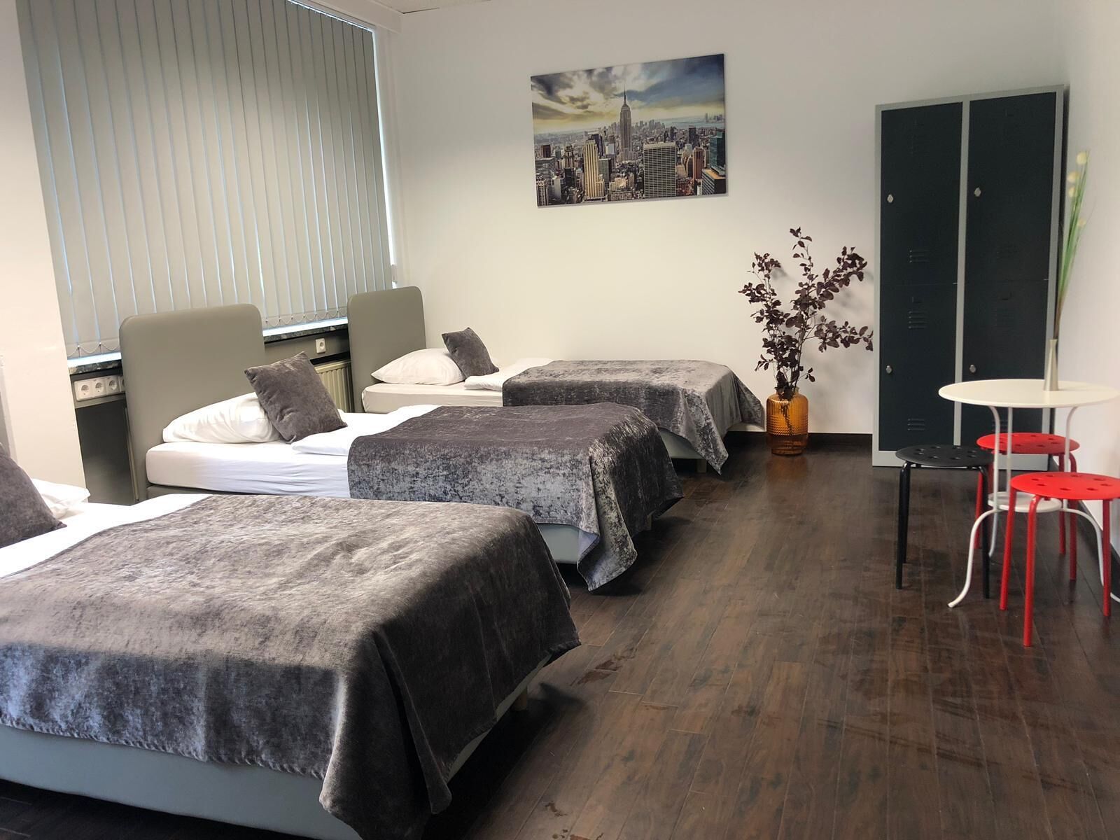 A hotel room with two beds, a small table, and a chair. There is a window with blinds, a painting on the wall, and a door with a lock.