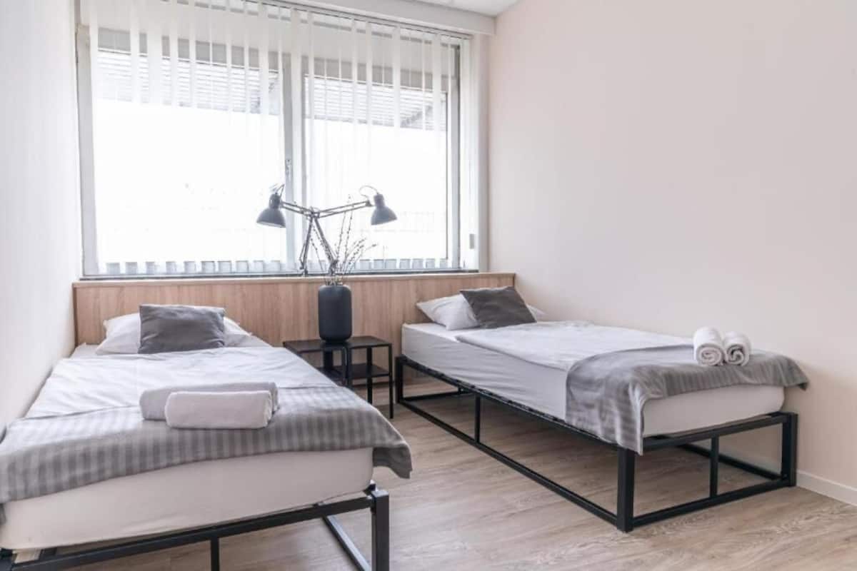 A room with two single beds, a window with blinds, a desk with a lamp, and a vase with branches.
