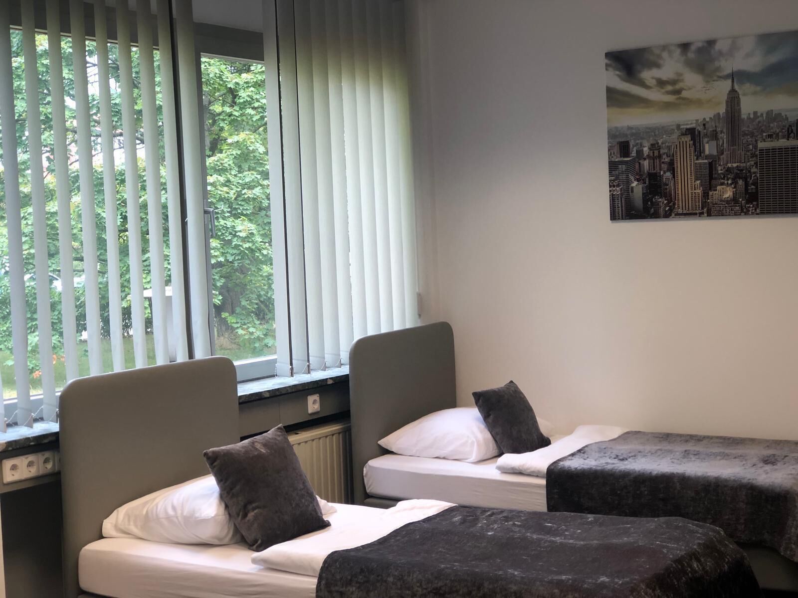 Ein Hotelzimmer mit zwei Betten, einem Stadtbild an der Wand und einem Fenster mit Jalousien.