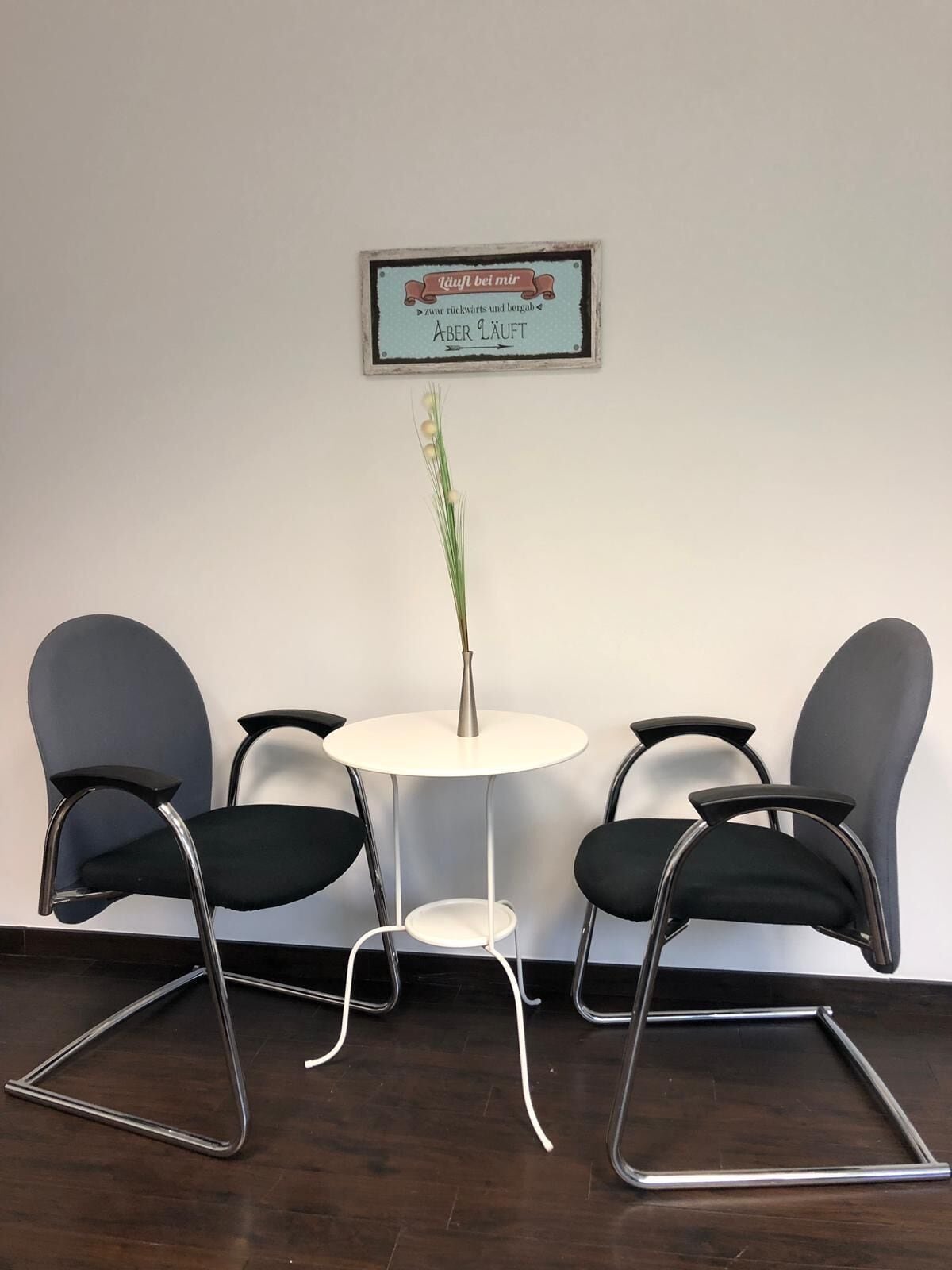 Uma pequena área de reuniões com uma mesa branca e quatro cadeiras pretas. Há um letreiro emoldurado acima da mesa e um vaso com plantas verdes sobre a mesa.