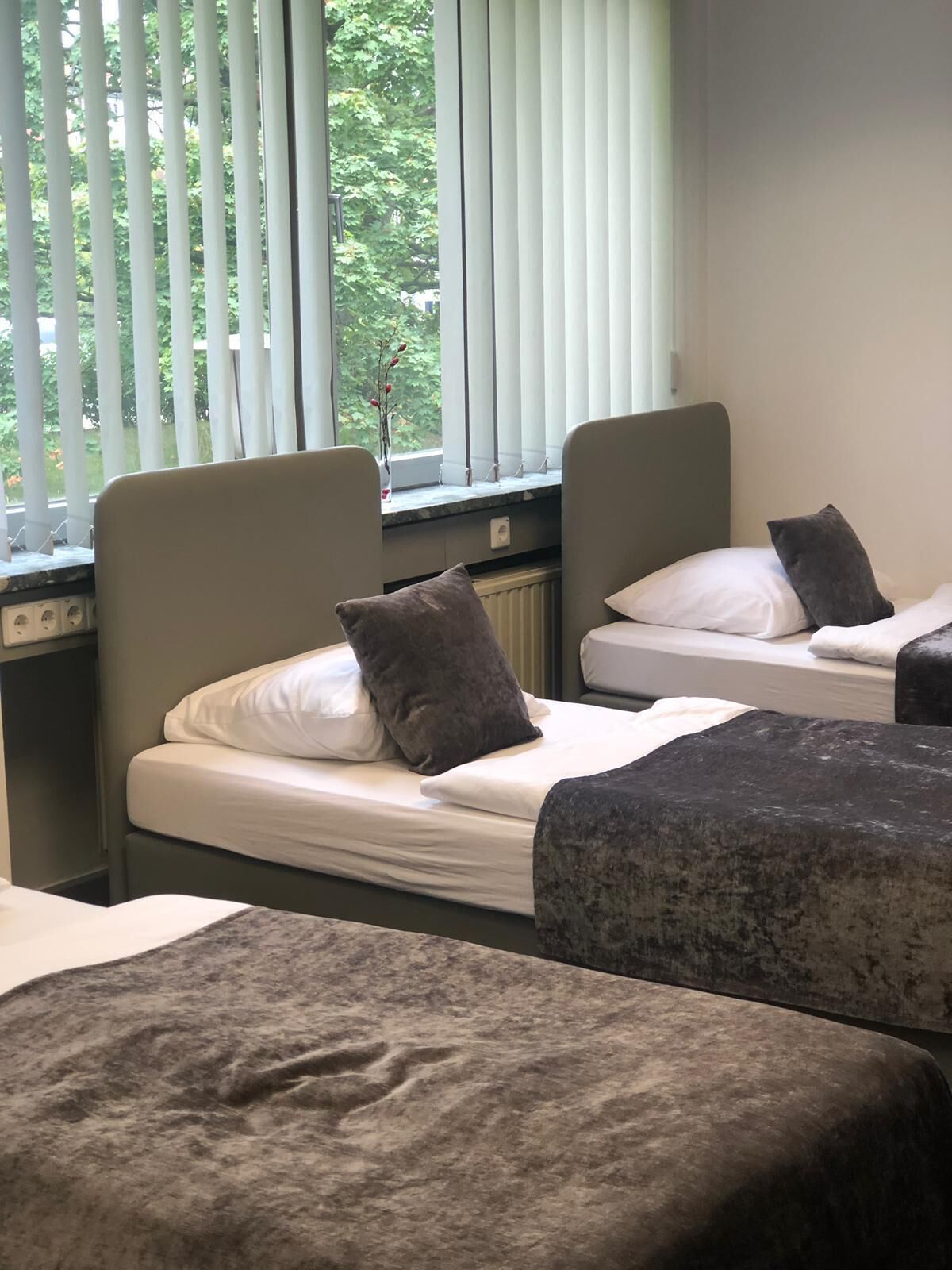 A room with two single beds, each with a pillow and a blanket. There is a window with blinds, and a radiator is visible on the wall.