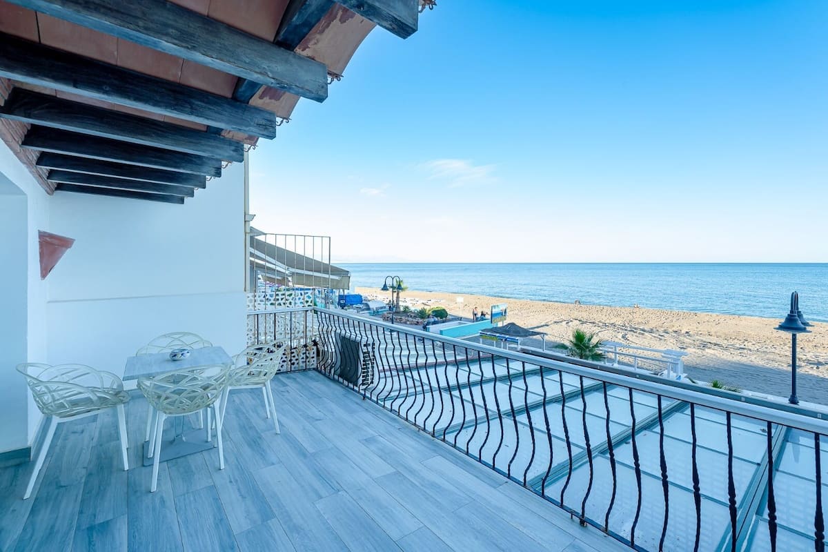 A balcony with a view of the beach, a pool, and outdoor furniture.