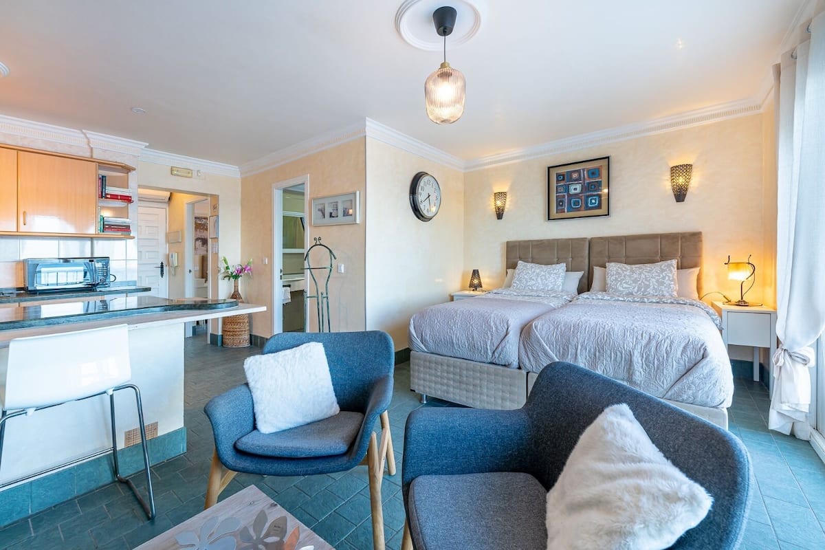 A modern hotel room with a kitchenette, two beds, a seating area with chairs, and a dining table.