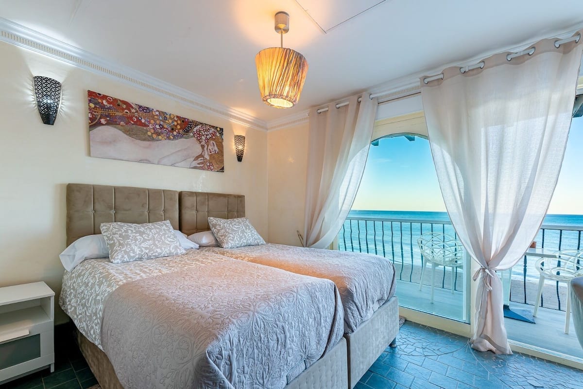 A double bed with a headboard, a bedside table, a nightstand, a painting, a chandelier, a balcony with a view of the sea, and sheer curtains.