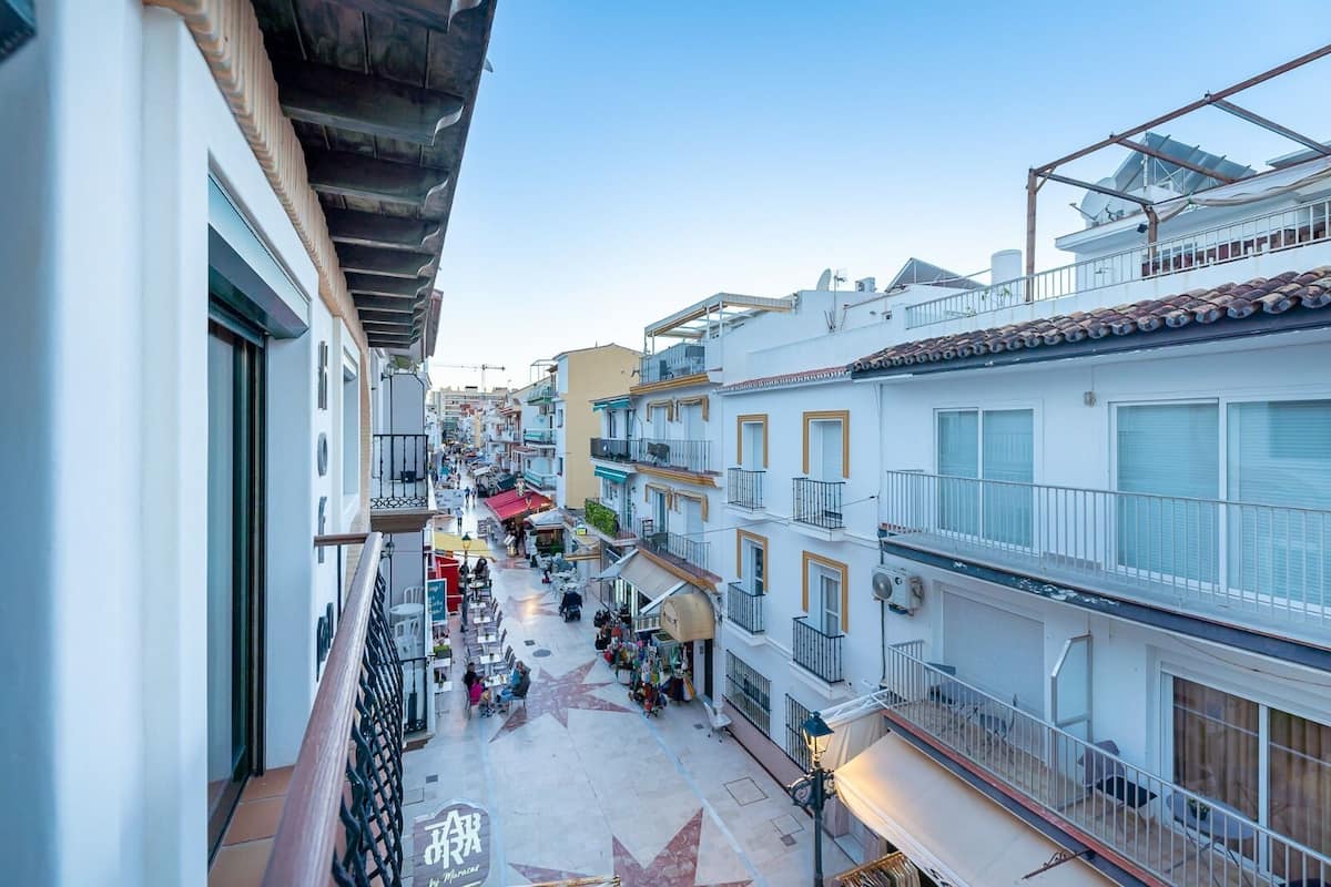 A narrow street with buildings on both sides, outdoor seating areas, and a clear sky.