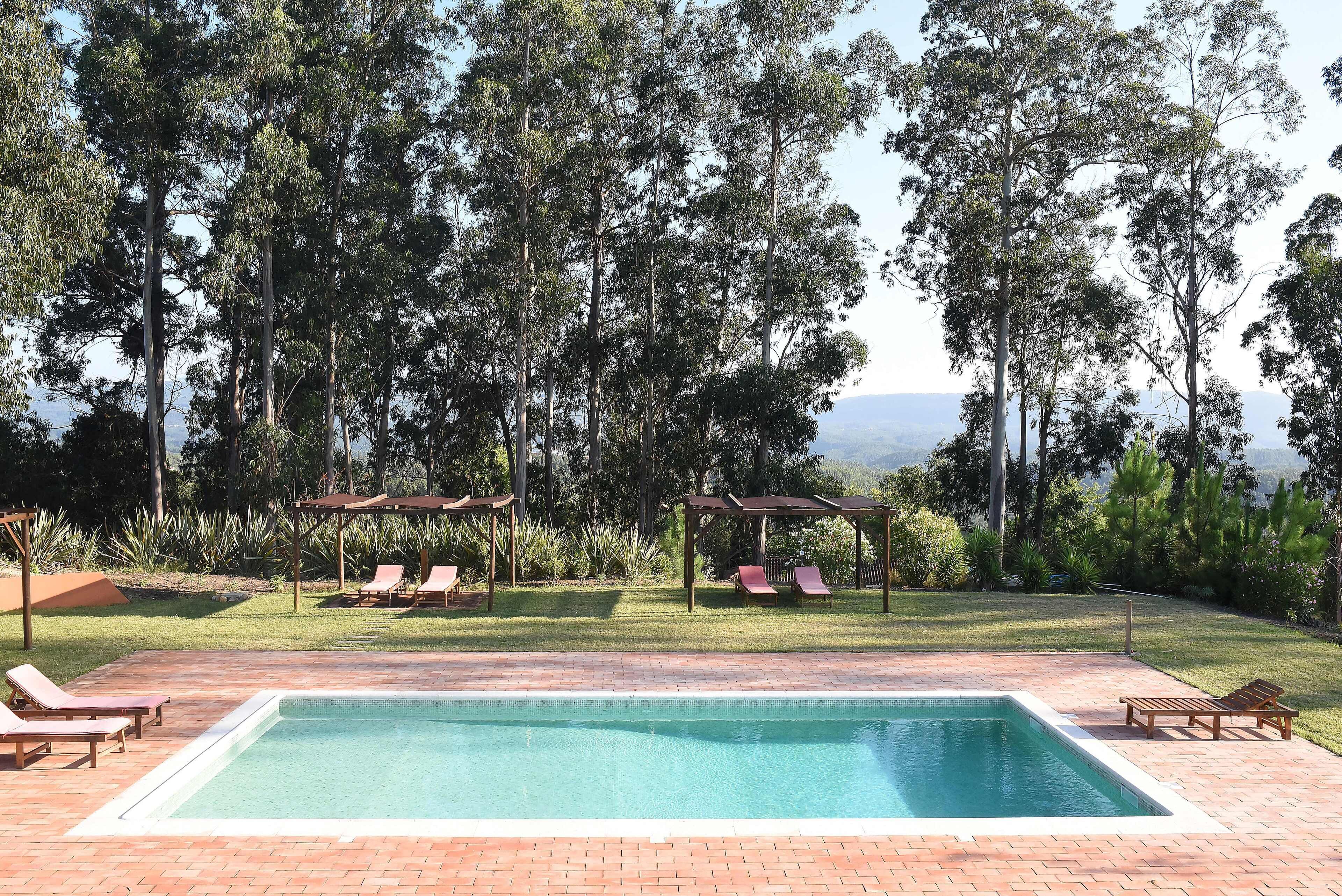Ein rechteckiger Pool mit klarer, blauer Wasserfläche, umgeben von einer Ziegelterrasse. Im Hintergrund sind Liegestühle und Holzpergolen mit roten Dächern zu sehen.