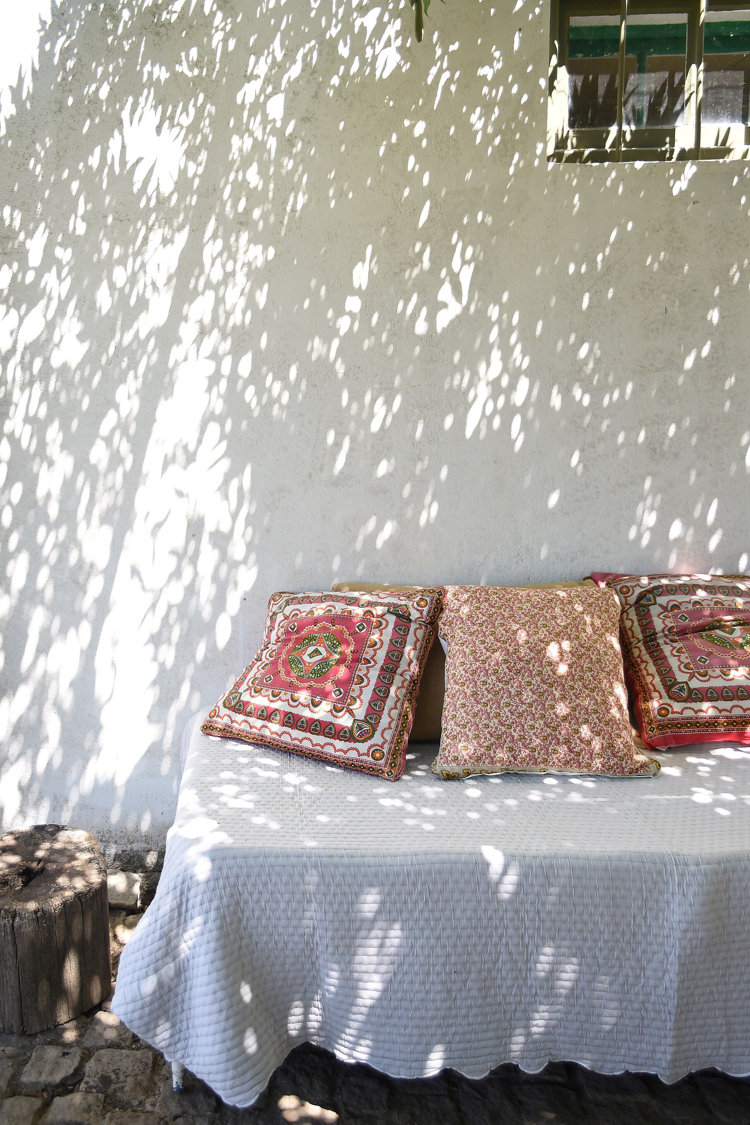 Cama com almofadas estampadas e colcha branca, diante de uma parede com janela e árvore projetando sombras.