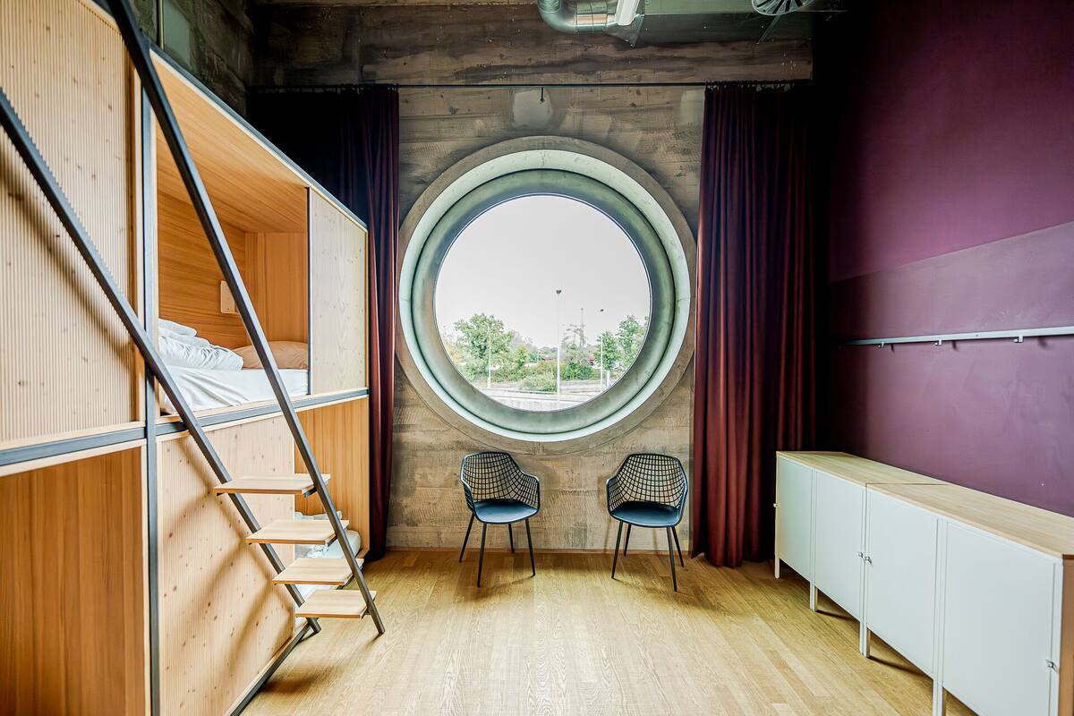 A room with a bunk bed, two chairs, a round window, and a cabinet.
