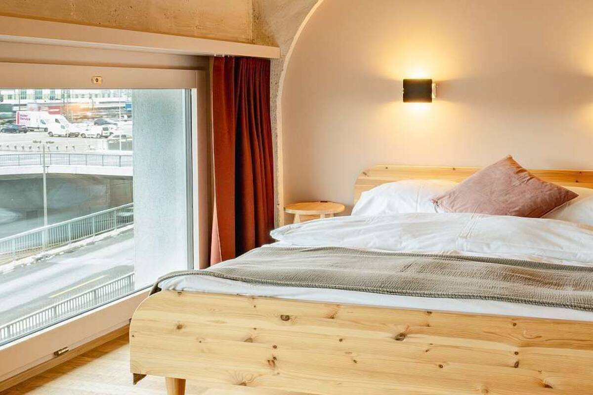 A bedroom with a wooden bed, two bedside tables, a window with a view of a road, and two wall-mounted lights.