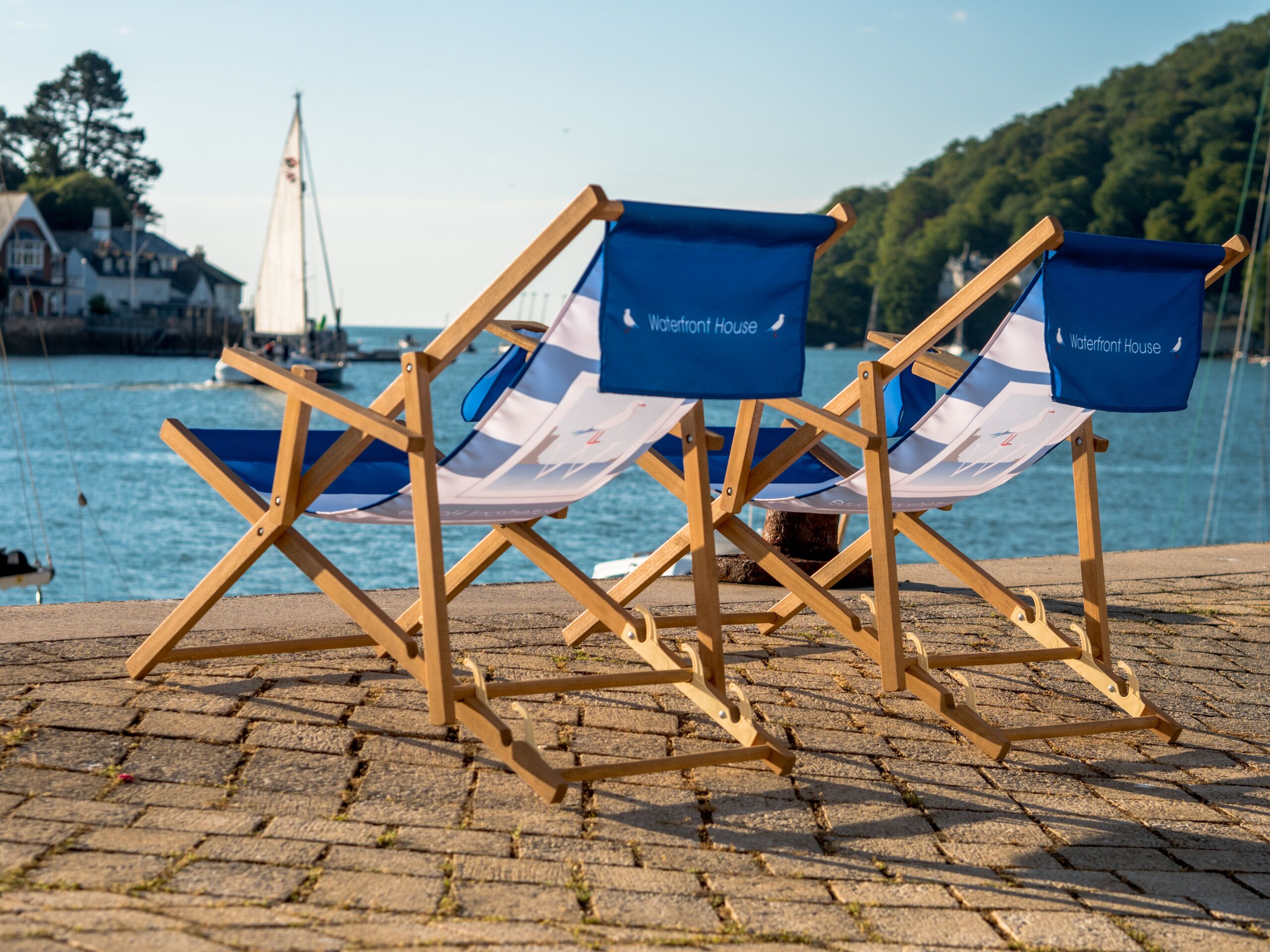 Two wooden folding chairs with blue and white striped fabric, labeled "Waterfront House," positioned on a paved surface by a body of water with a sailboat and buildings in the background.
