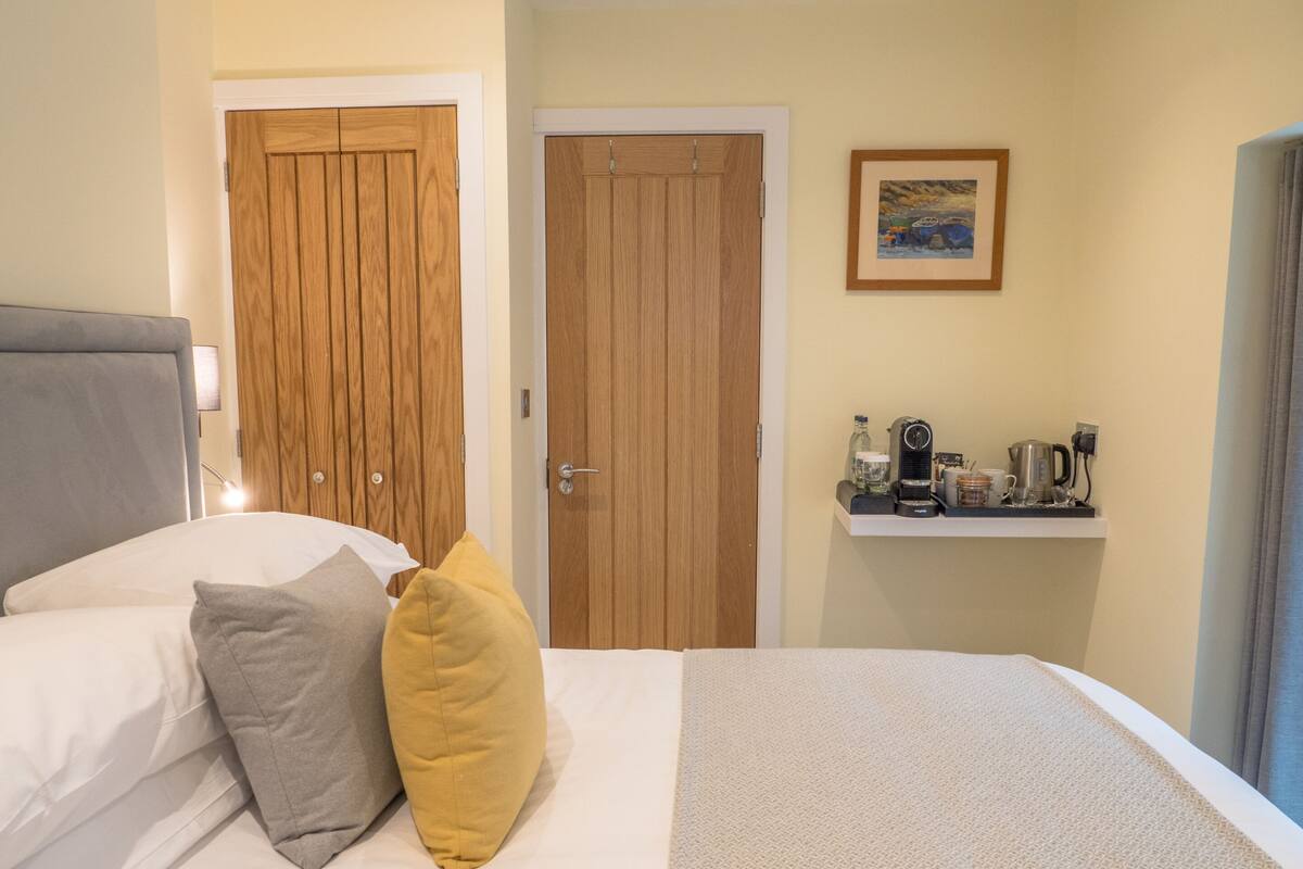 Ein Hotelzimmer mit einem Bett, zwei Holztüren, einem Regal mit einer Kaffeemaschine und einem gerahmten Bild an der Wand.