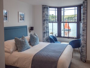 A hotel room with a large bed, two chairs, a window with a view of a body of water and a sailboat, and a headboard with a blue panel. - Waterfront House (Dartmouth)