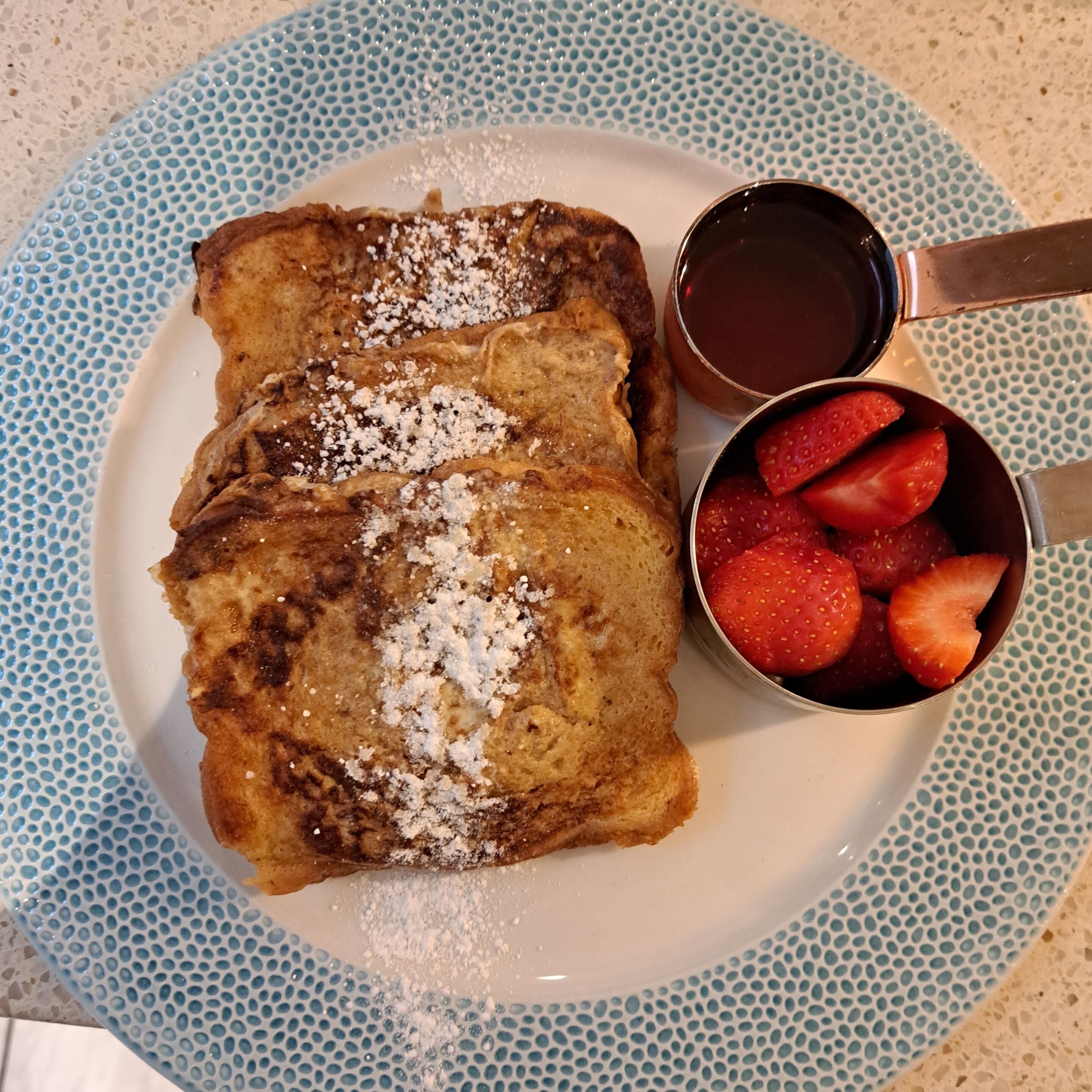 A plate with two pieces of food, possibly pancakes, topped with powdered sugar and a small bowl of strawberries.