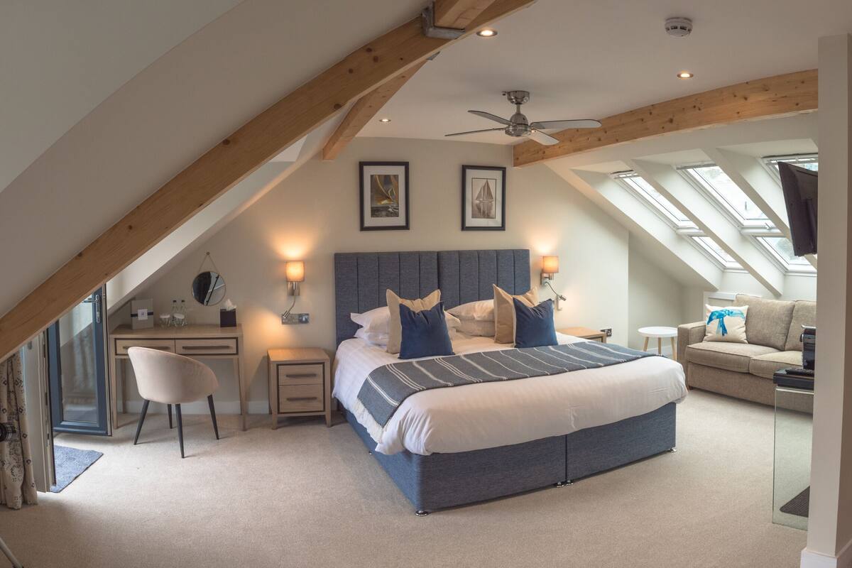 Ein Schlafzimmer mit einem großen Bett, einem Schreibtisch, einem Sessel und einem Sofa. Es gibt Dachfenster und einen Deckenventilator.