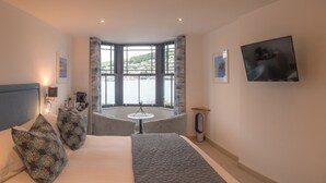 Ein Hotelzimmer mit einem großen Bett, einem Schreibtisch, einem Sessel, einem kleinen Tisch, einem Fernseher und einem Erkerfenster mit Meerblick.