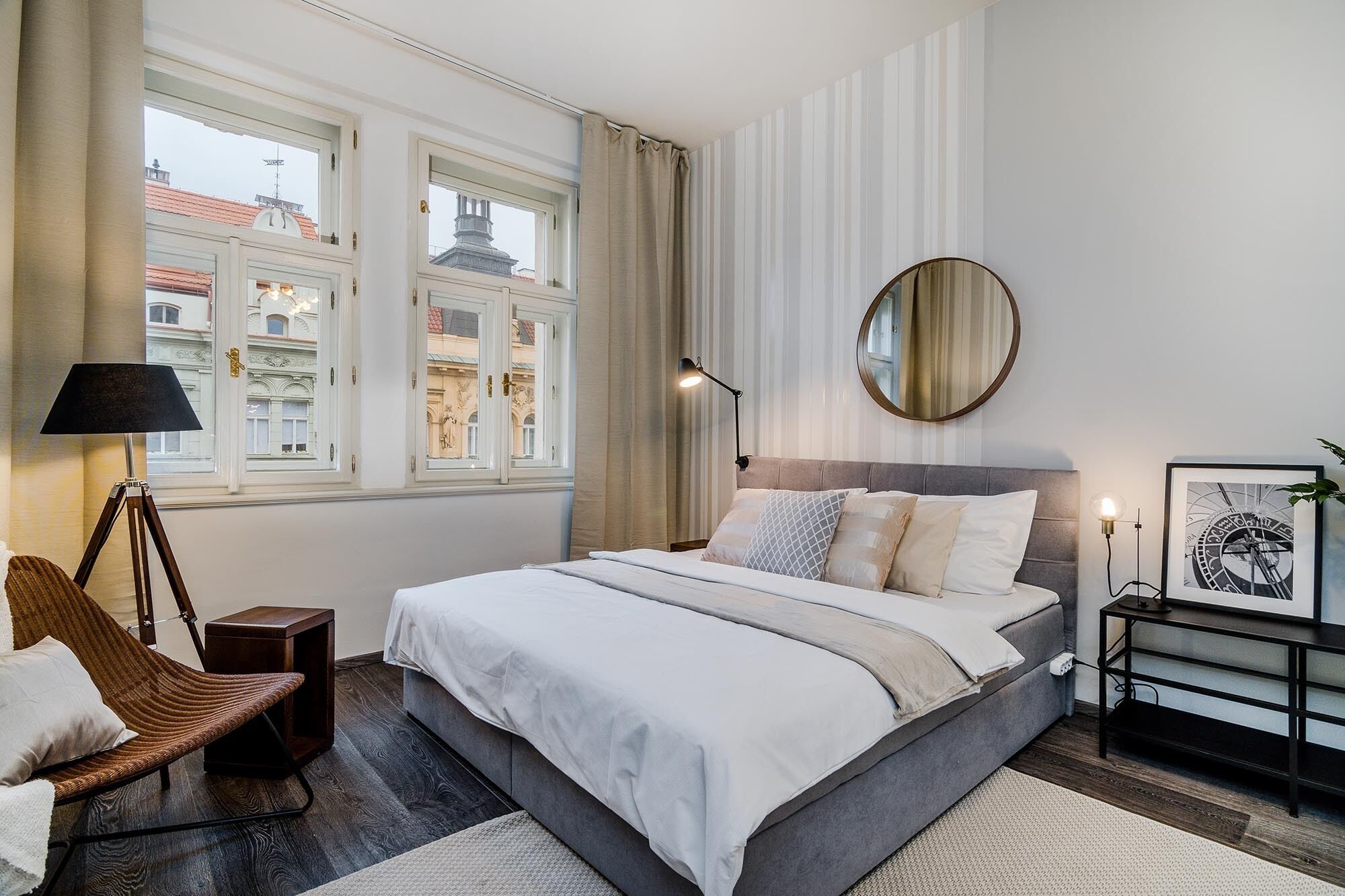 A modern bedroom with a large bed, a round mirror, a side table, and a view of buildings through the window.