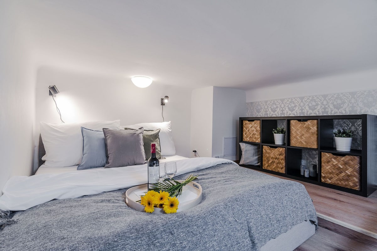 Ein modernes Schlafzimmer mit Bett, Nachttischlampen, einem Nachttisch mit Flasche und Gläsern, einem Tablett mit Blumen und einer dekorativen Wandeinrichtung mit Pflanzen.