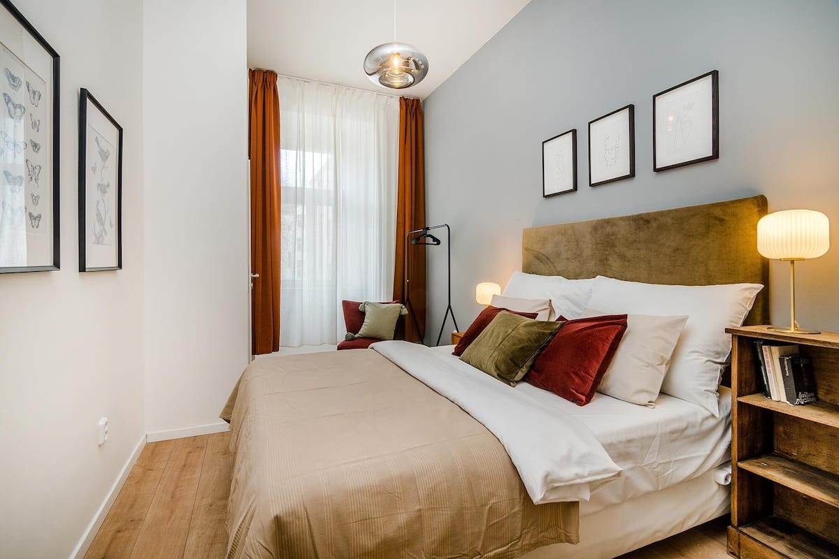Ein ordentlich eingerichtetes Schlafzimmer mit einem Bett, Nachttischlampen, einem Nachttisch mit Büchern und gerahmten Kunstwerken an der Wand.